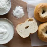 An overhead view of the three ingredients for bagels—flour, Greek yogurt, and baking powder—next to freshly baked 3-ingredient bagels on a wooden board.