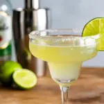 A perfectly crafted margarita representing the best margarita formula, with a salted rim, lime garnish, and ingredients like tequila and a shaker in the background.