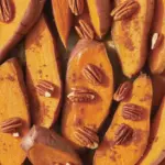A close-up of a candied sweet potatoes recipe, featuring caramelized sweet potato slices in a brown sugar glaze, topped with toasted pecans in a baking dish.