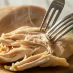 A close-up shot of two forks shredding a juicy, moist chicken breast, highlighting the key technique for making perfect moist shredded chicken.