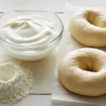 Ingredients for making bagels with normal yogurt, including a bowl of yogurt, flour, and raw bagel dough on a wooden table.