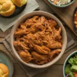 A rustic wooden table displays a central bowl of pulled chicken surrounded by perfect side dishes like coleslaw, cornbread, and a fresh salad.